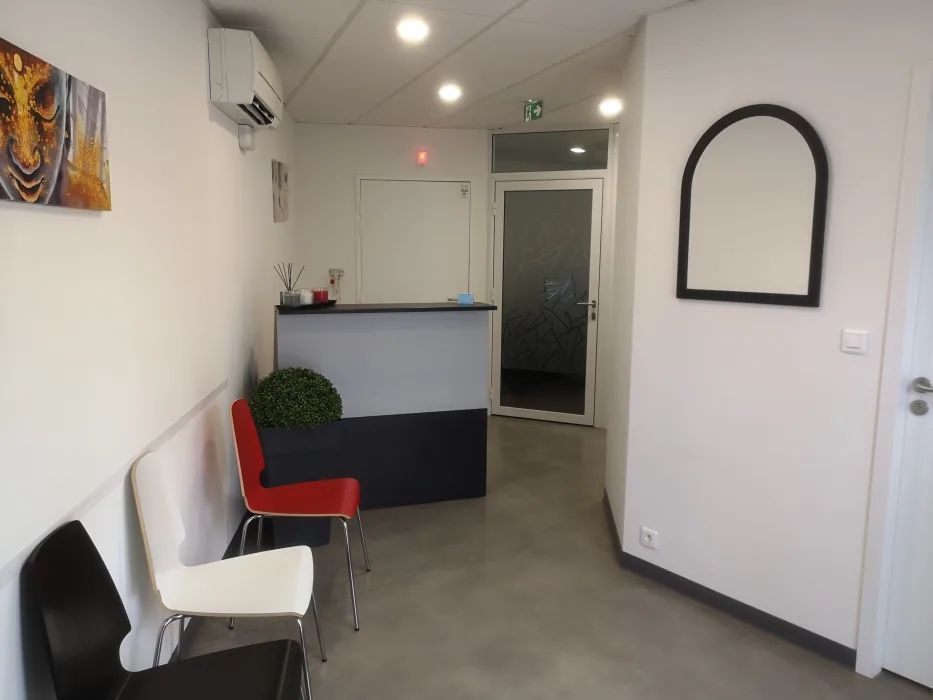 Petite salle d'attente avec chaises rouges et blanches, comptoir d'accueil et décorations murales dans un couloir blanc.