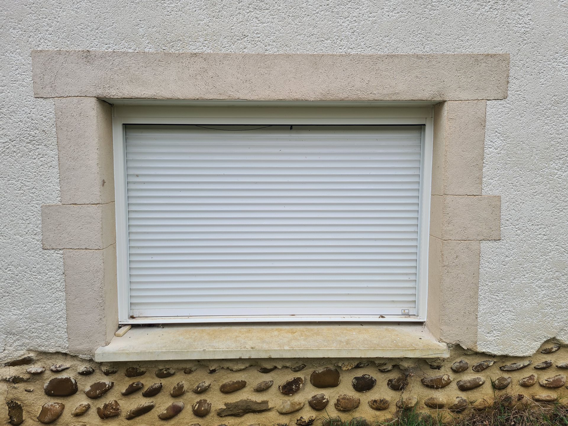 Volet de fenêtre blanc fermé, encastré dans un mur en stuc beige au-dessus d'une bordure de galets.