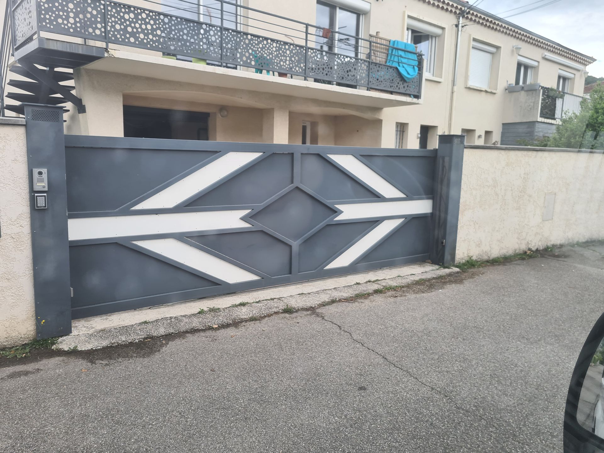 Entrée d'une maison moderne avec un portail géométrique gris foncé et un mur blanc, donnant sur une rue calme.