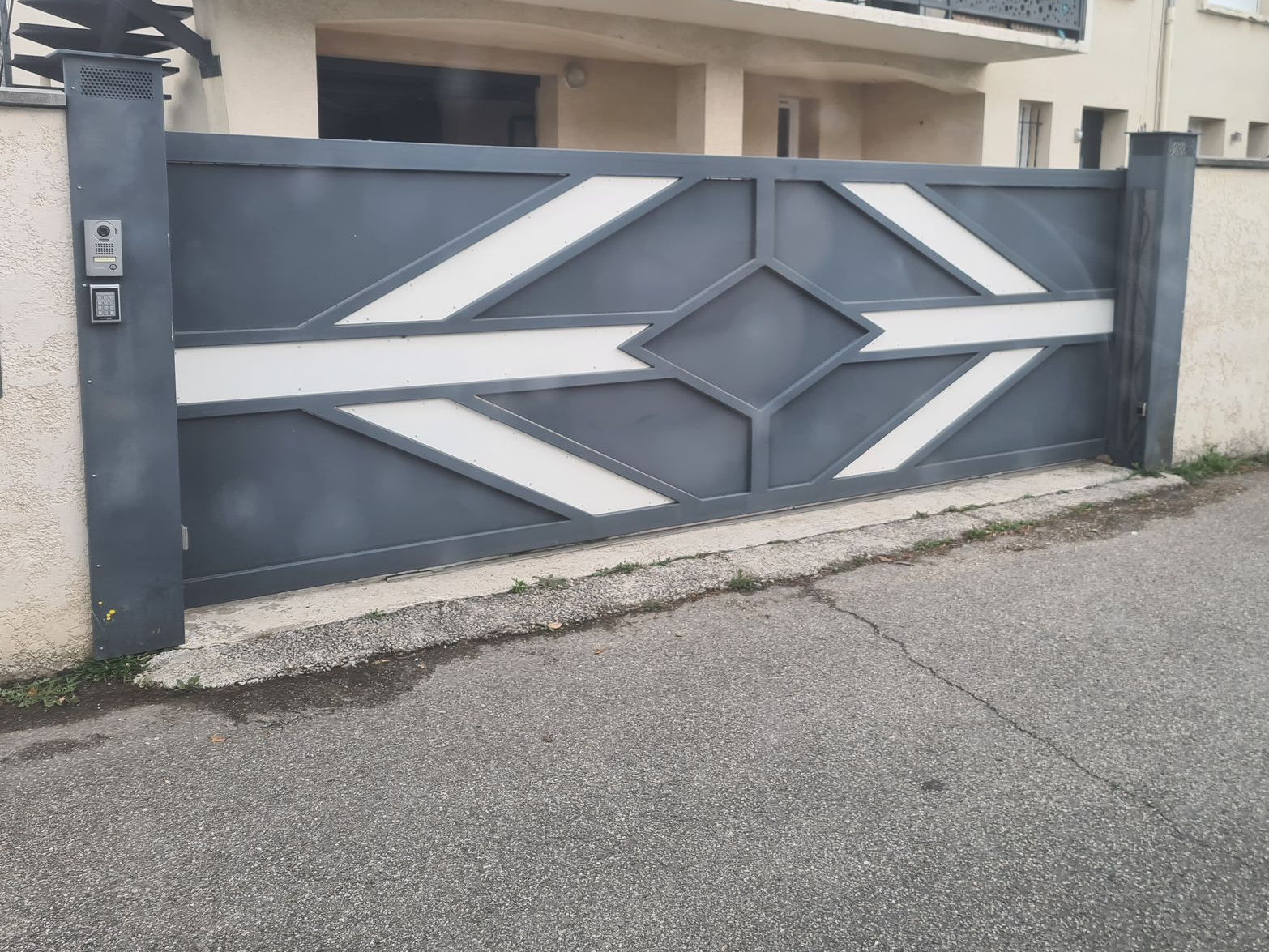 Portail d'allée en métal gris avec panneaux géométriques blancs devant une maison et la rue.