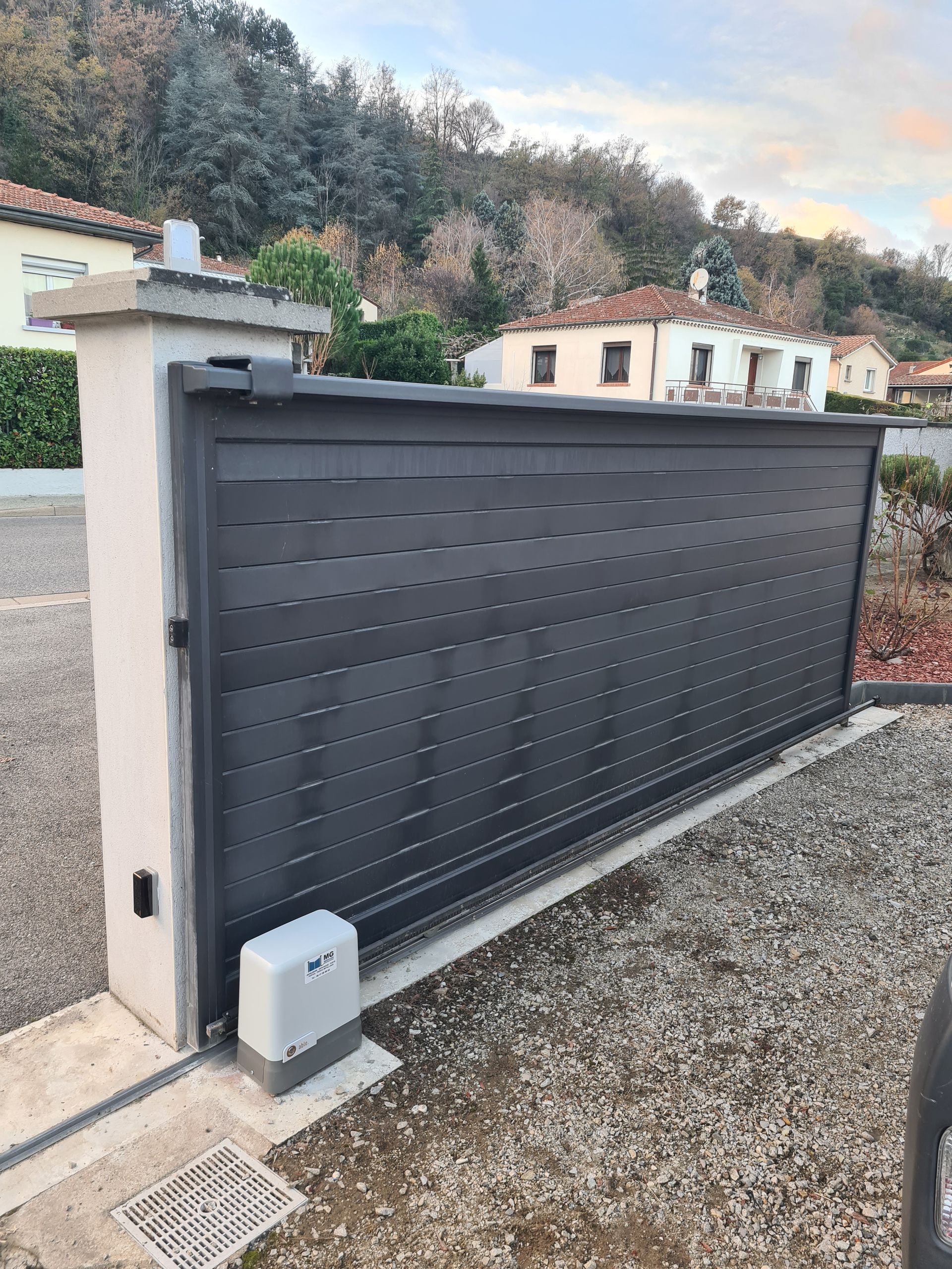 Portail coulissant gris foncé donnant sur une allée, à côté d'un pilier en stuc, avec un petit boîtier de commande et du gravier au premier plan.