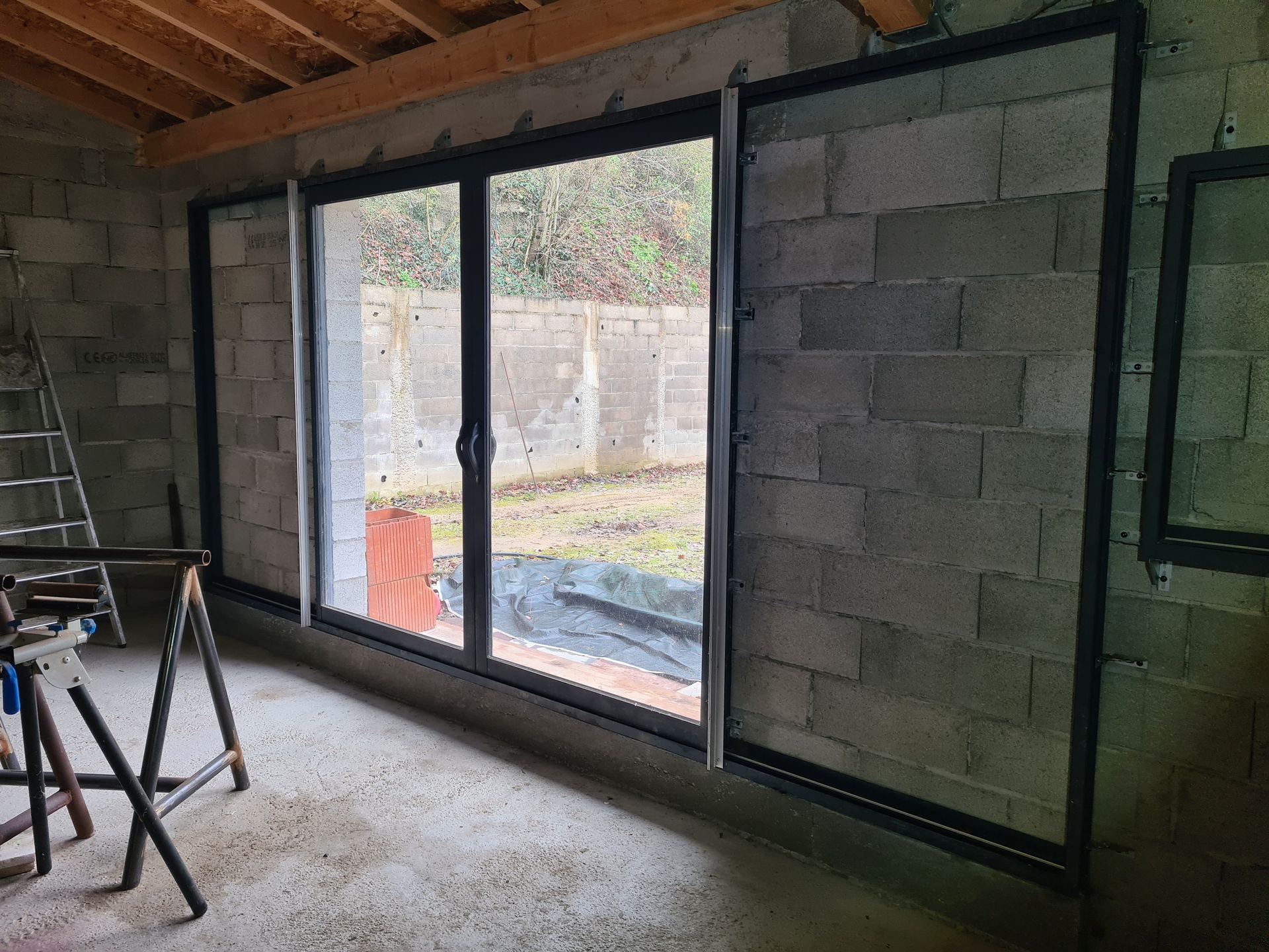 Pièce inachevée avec de grandes portes coulissantes en verre à cadre noir donnant sur une terrasse et un jardin.