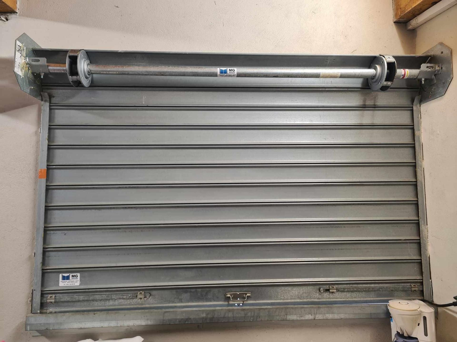 Volet roulant métallique argenté fermé, avec un mécanisme à barre horizontale au-dessus.