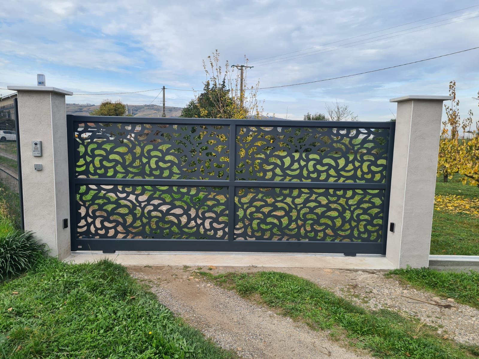 Portail d'allée décoratif en métal noir entre des piliers blancs, avec un chemin gazonné devant et un ciel nuageux derrière.