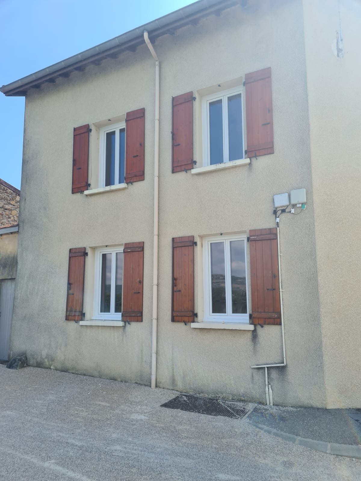 Maison beige à deux étages avec volets rouges et fenêtres à cadre blanc, située dans une rue pavée.