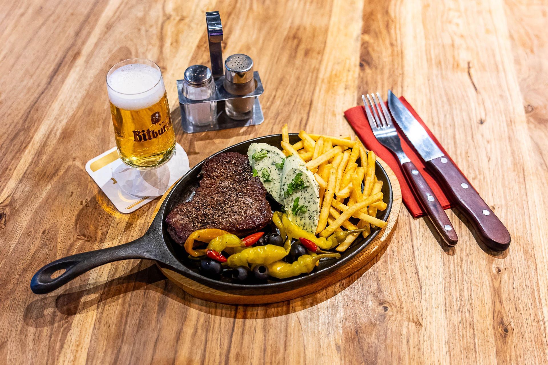 Eine Pfanne gefüllt mit Steak und Pommes Frites auf einem Holztisch neben einem Glas Bier.