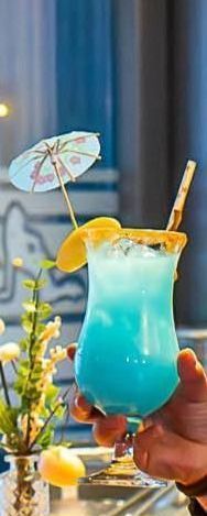 Eine Hand hält einen blauen Cocktail mit Sonnenschirm, Orangenscheibe und Strohhalm.