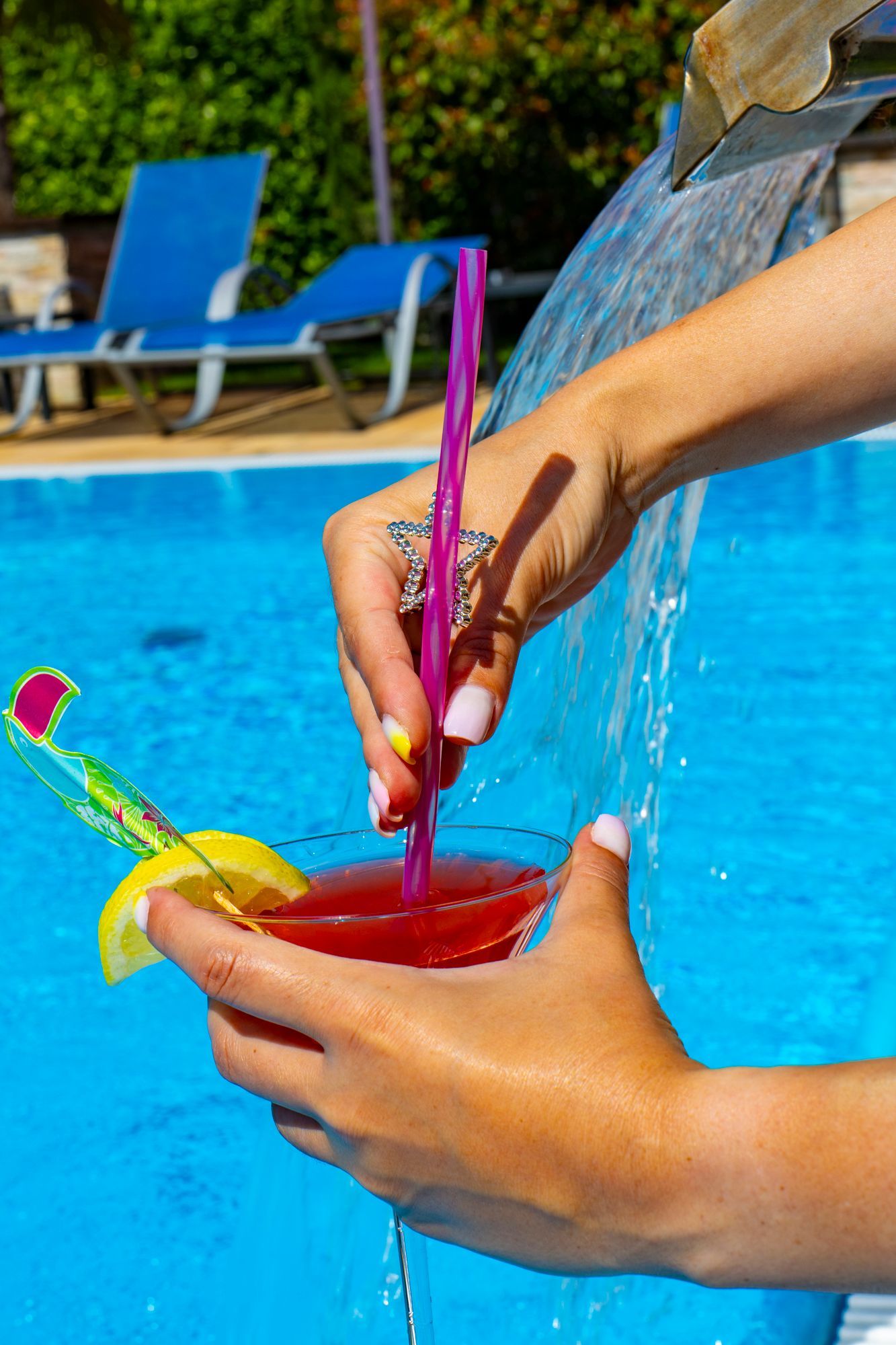Person gießt Wasser in einen roten Cocktail am Pool. Der Cocktail ist mit einem Strohhalm und Früchten garniert; im Hintergrund Liegestühle.