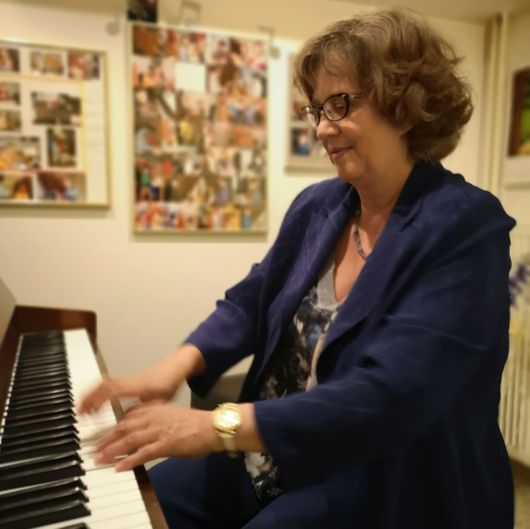 Eine Frau spielt Klavier in einem Zimmer mit Fotos an der Wand. Sie trägt eine Brille und eine blaue Jacke.