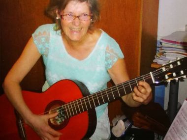 Frau spielt eine rote Akustikgitarre, lächelt drinnen, trägt eine Brille und ein blaues Hemd.