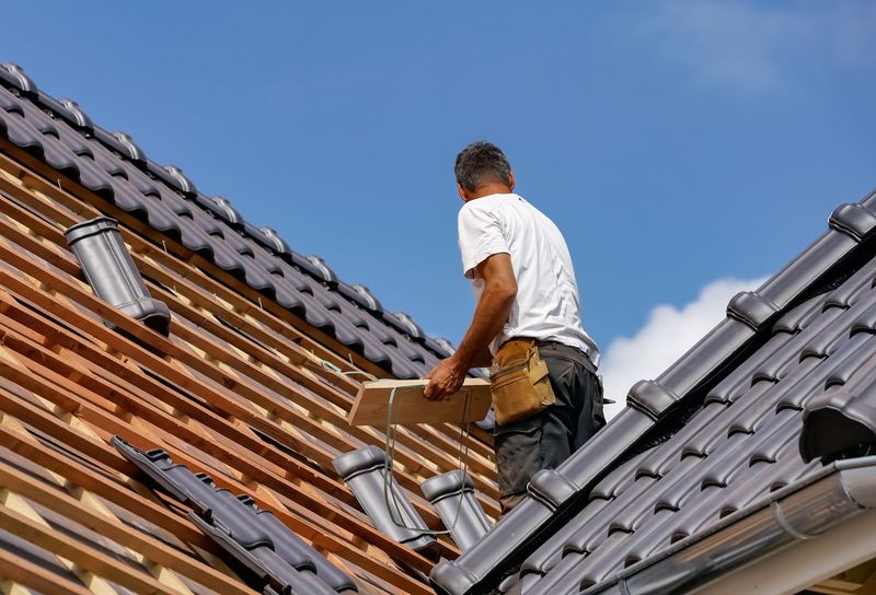 Une personne en chemise blanche et ceinture à outils travaille sur le toit d'une maison, installant des tuiles foncées sur des liteaux en bois.