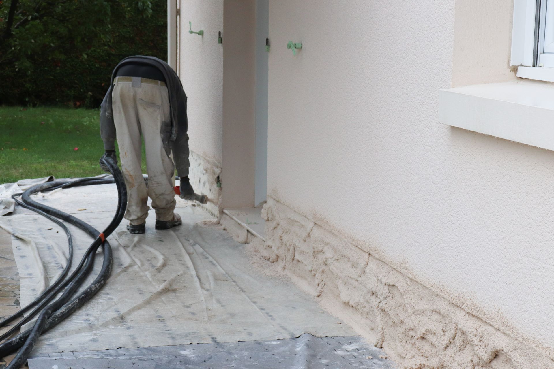 Une personne utilise un tuyau d'arrosage pour appliquer un enduit ou un mastic de couleur claire à la base d'un mur extérieur.