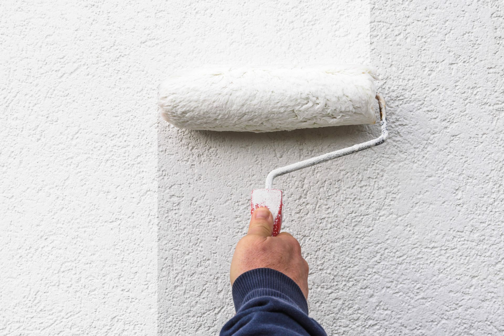 Une main tenant un rouleau à peinture applique de la peinture blanche fraîche sur un mur texturé de couleur claire.