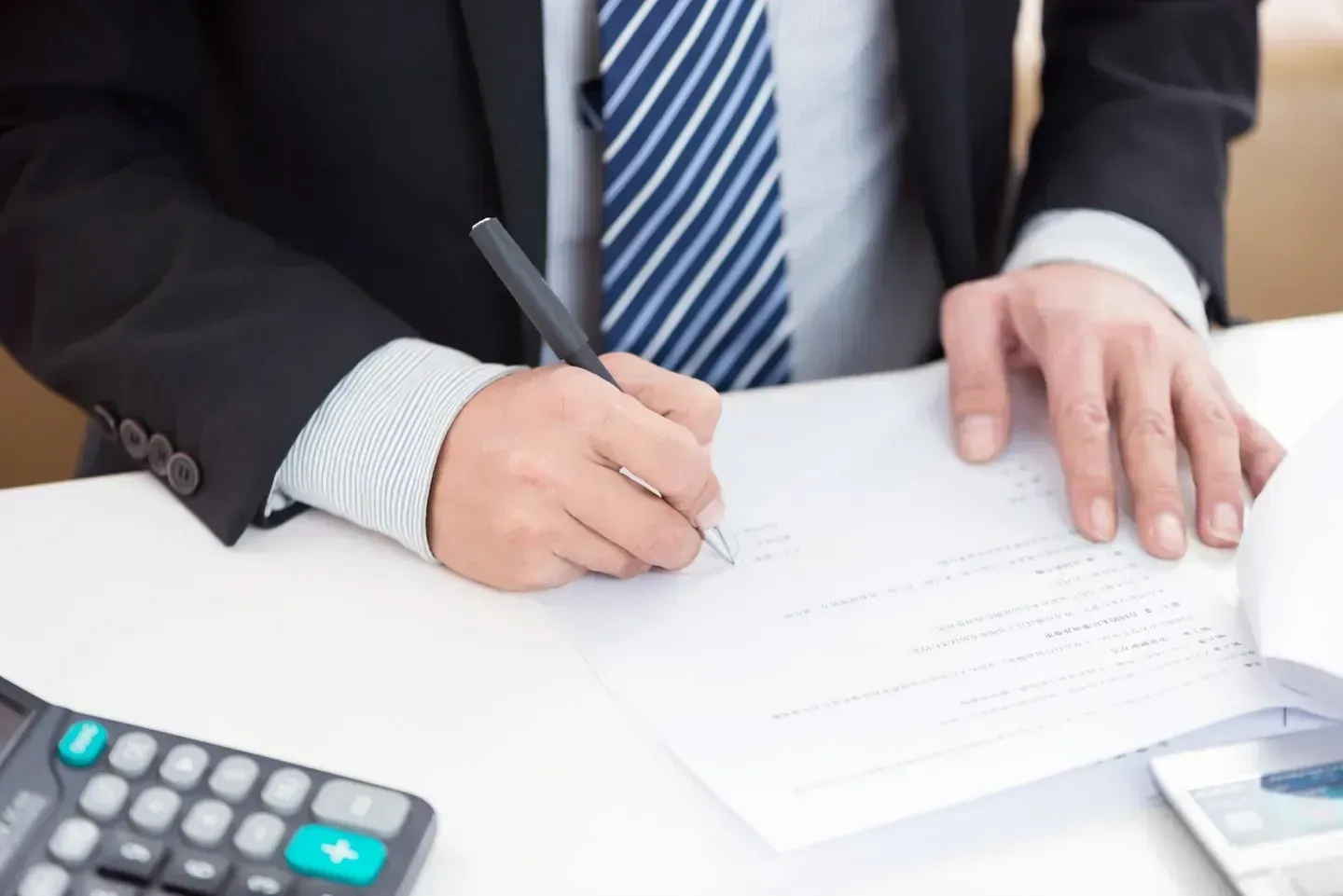 Persona con traje firmando un documento en un escritorio, con calculadora visible, entorno de oficina.