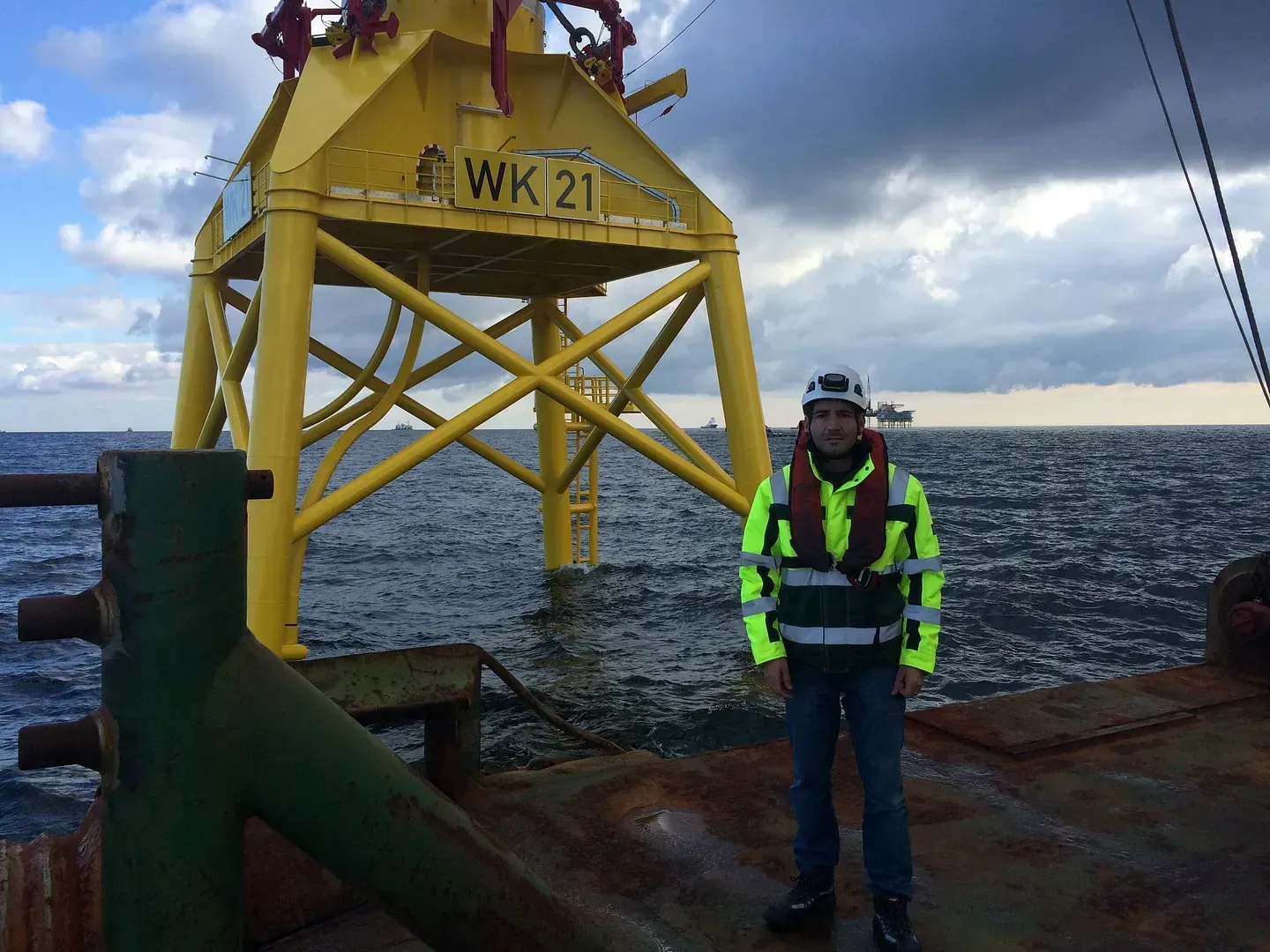 Persona con equipo de seguridad se encuentra en una plataforma cerca de una estructura amarilla en alta mar, mar, cielo nublado.
