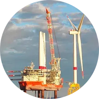 Aerogenerador marino en construcción mediante un barco grúa rojo y blanco, con fondo de cielo nublado.