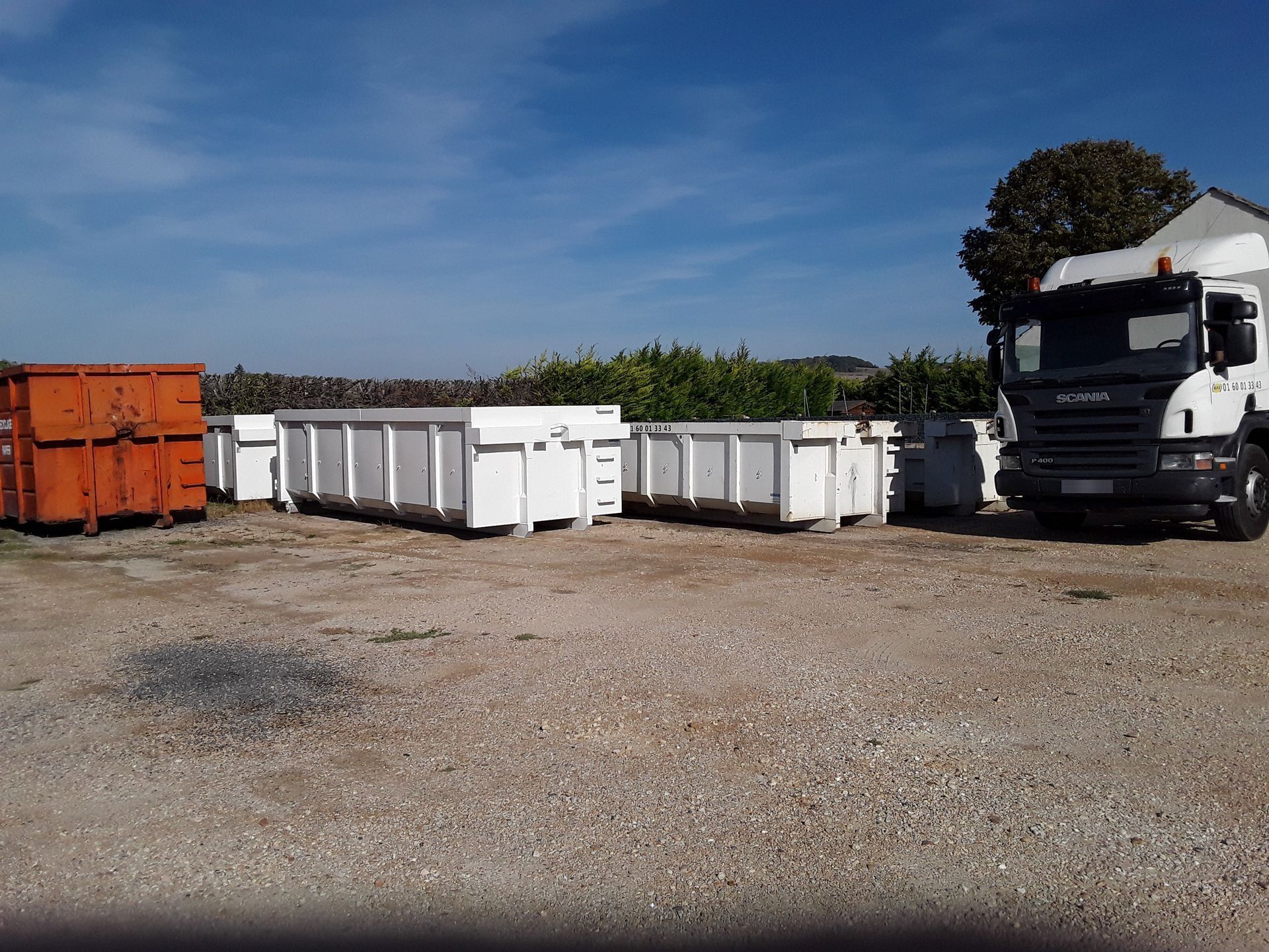 Des bennes à ordures sur un terrain gravillonné, à côté d'un camion blanc.