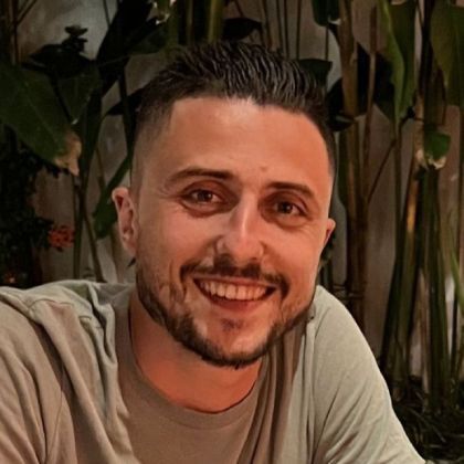 Un homme avec une barbe sourit à la caméra devant une plante.