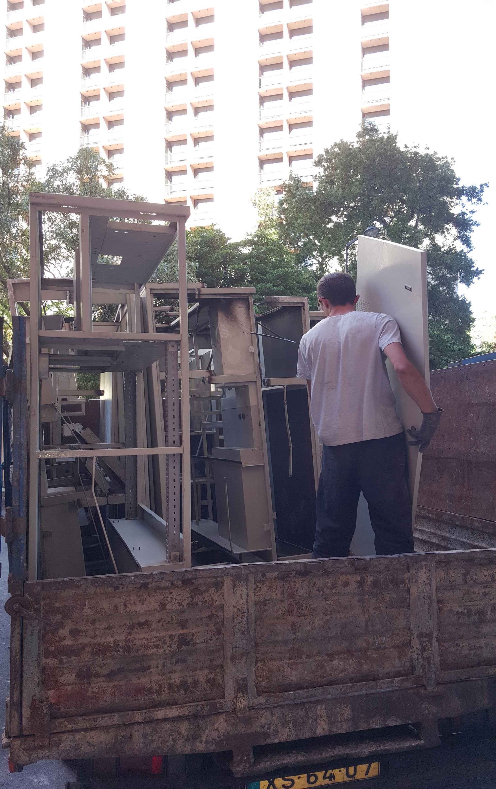 Homem carregando equipamento de metal em um caminhão enferrujado, em frente a um prédio alto.