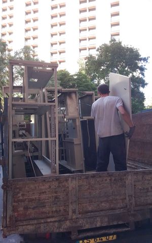 Homem carregando equipamento de metal em um caminhão enferrujado, em frente a um prédio alto.