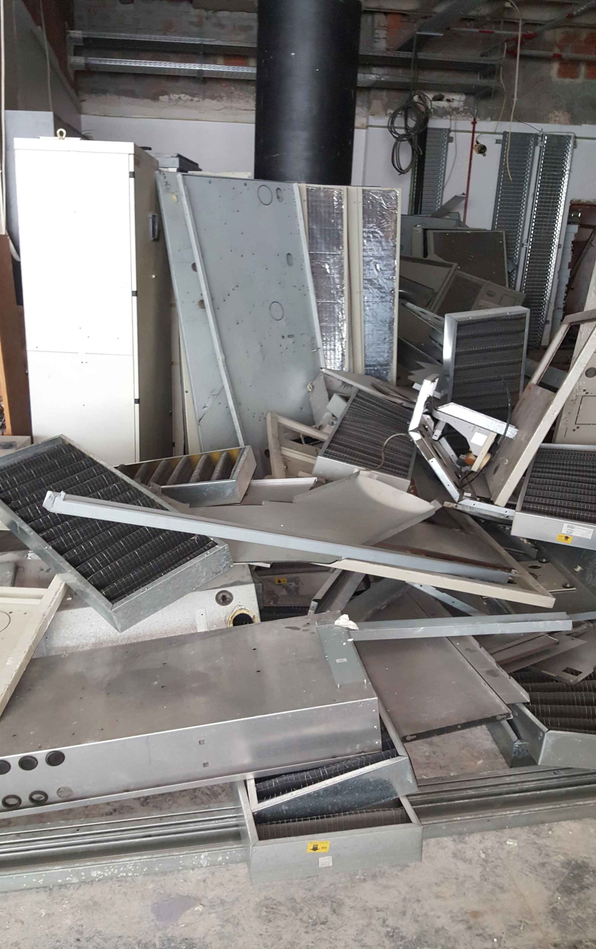 Pilha de sucata: chapas de metal dobradas, serpentinas de ar condicionado, grades e dutos em um piso de concreto.