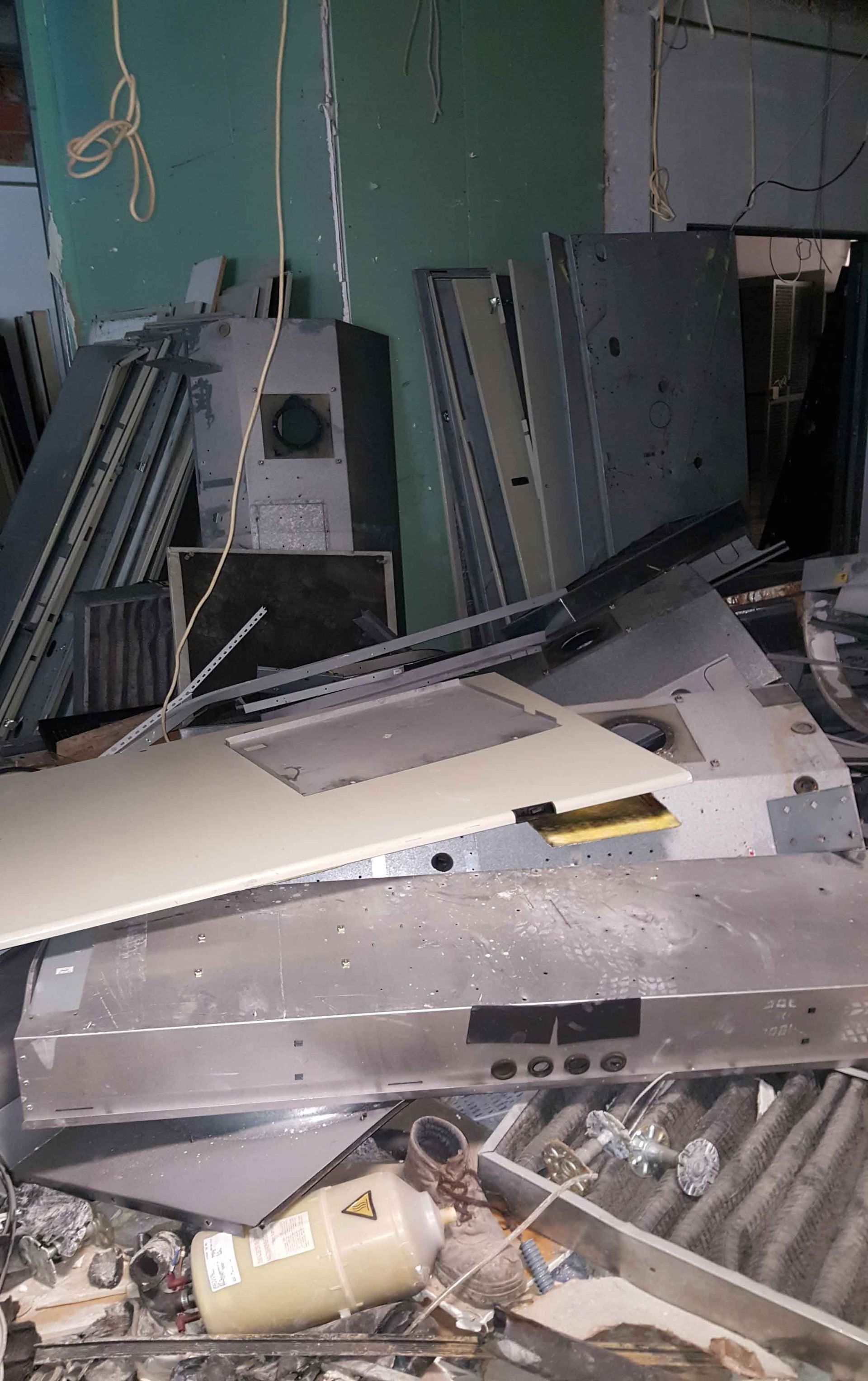 Pilha de entulho de metal e equipamentos em um ambiente industrial, com um cilindro branco e uma parede verde visíveis.