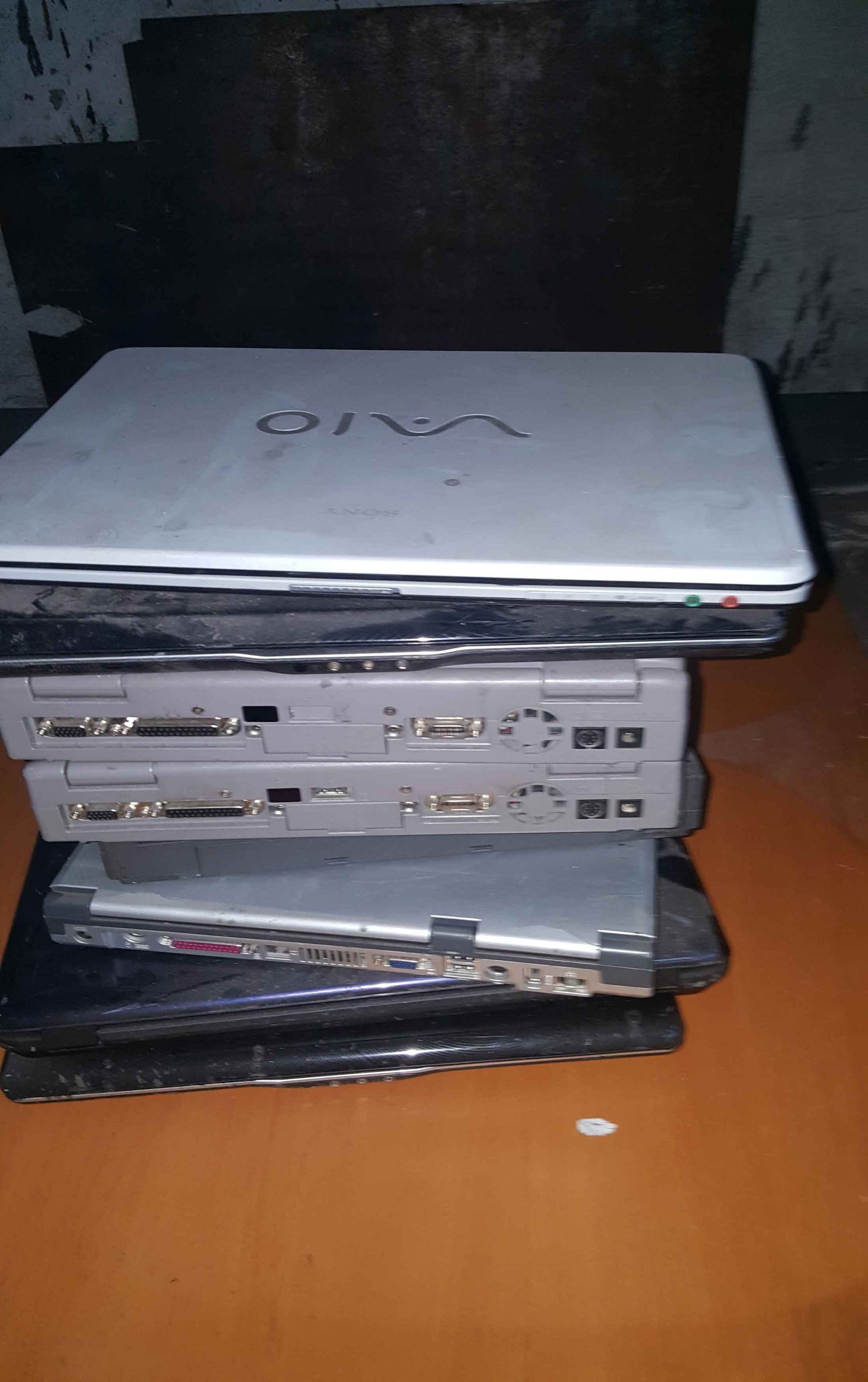 Pilha de vários laptops antigos, alguns prateados, outros pretos, sobre uma superfície marrom.