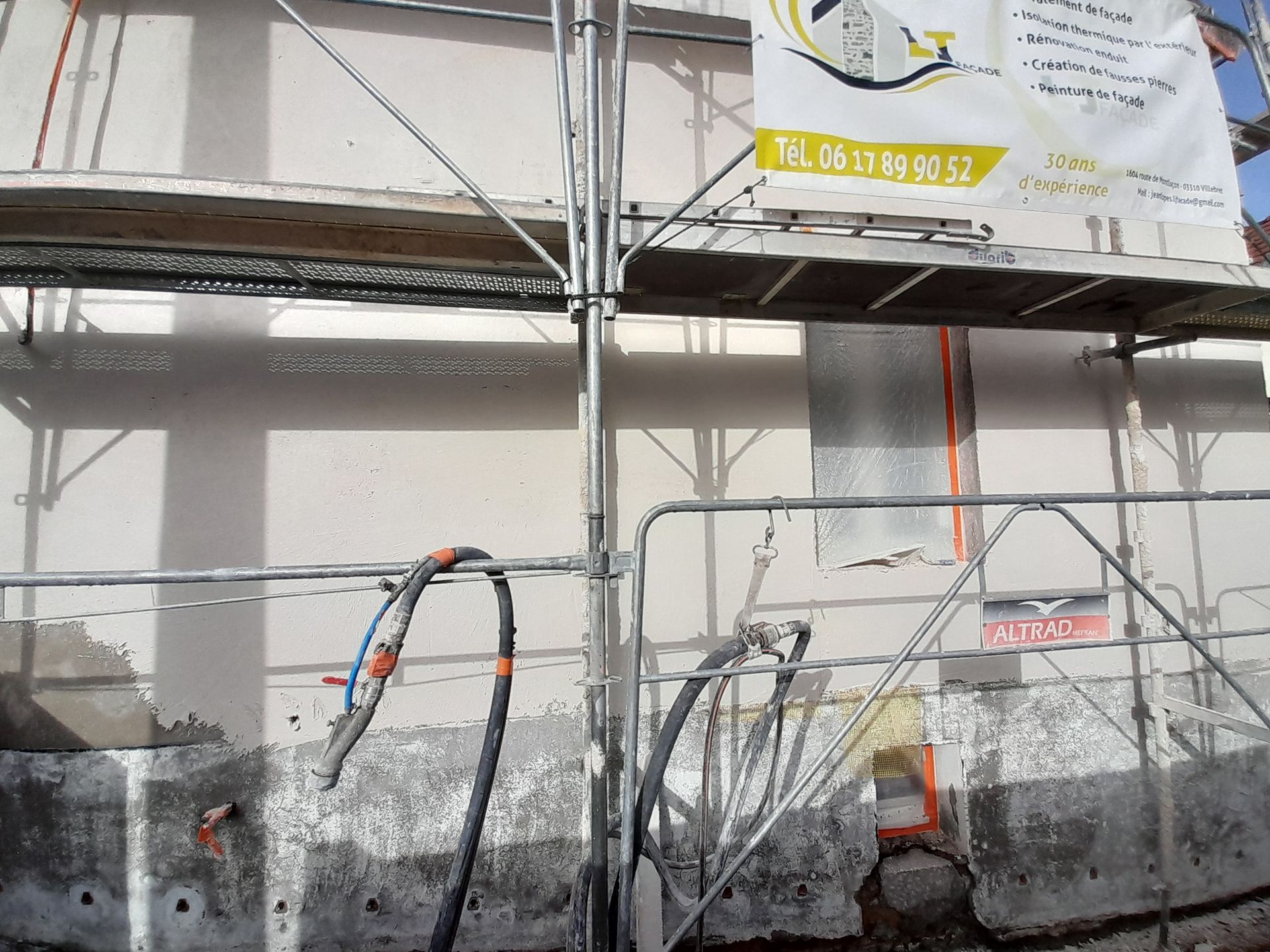 Deux ouvriers sur un échafaudage enduisent le mur extérieur d'un bâtiment ; l'un applique le plâtre, l'autre travaille en dessous.