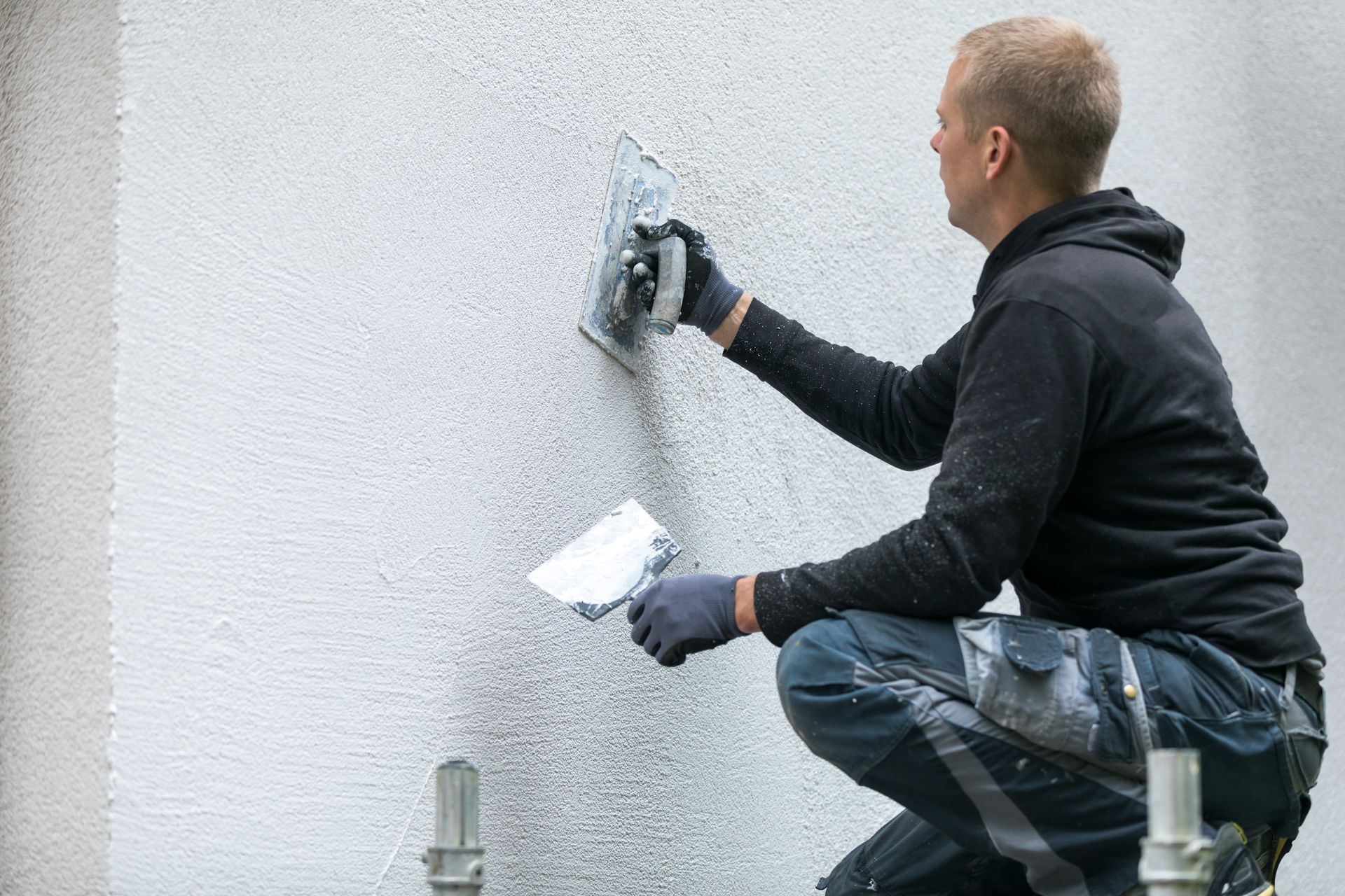 Un homme applique du stuc sur un mur extérieur à l'aide d'une truelle, portant des gants et un sweat-shirt à capuche.