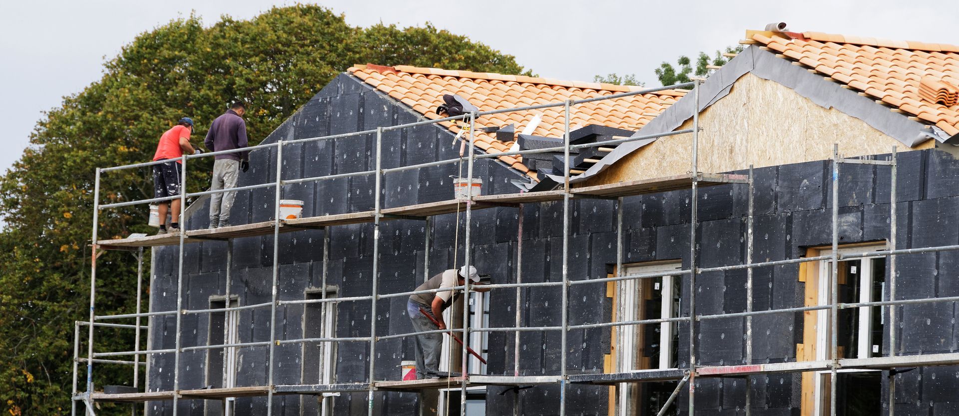 Des ouvriers du bâtiment travaillent sur un échafaudage en train de construire une maison. La maison présente des poutres apparentes et des panneaux d'isolation.