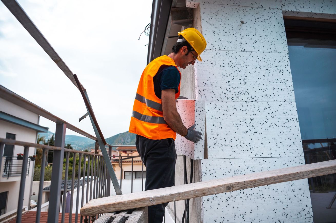 Ouvrier installant de l'isolant sur la façade d'un bâtiment, portant un gilet de sécurité et un casque de chantier.