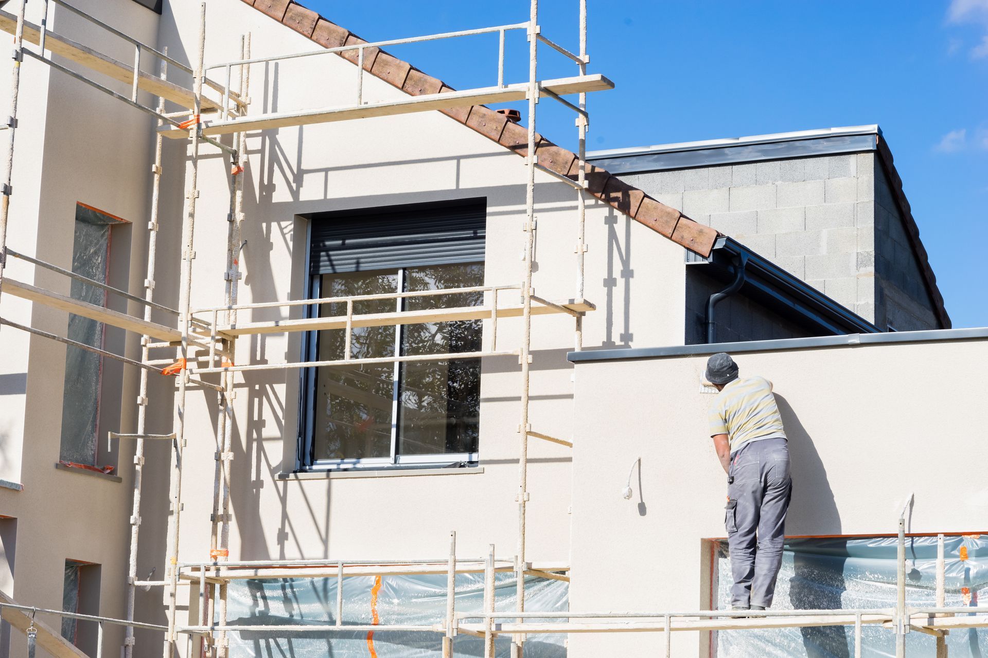 Un ouvrier enduit de plâtre le mur extérieur d'un bâtiment ; échafaudage en place ; journée ensoleillée.