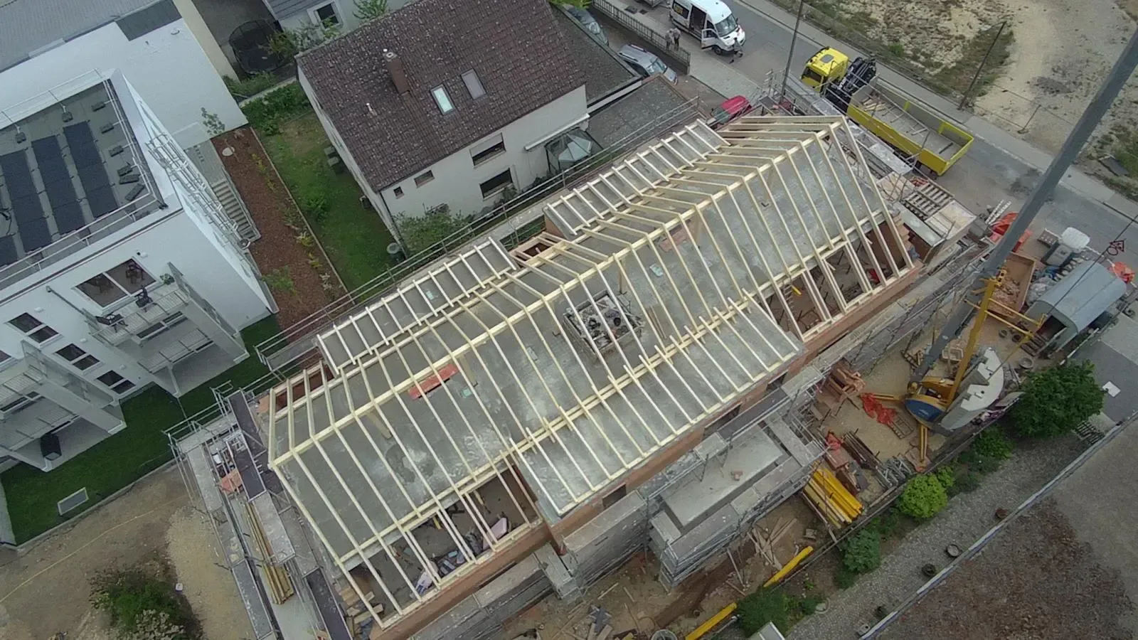 Luftaufnahme eines im Bau befindlichen Hauses mit freiliegendem Dachstuhl und in der Nähe befindlichen Baumaschinen.