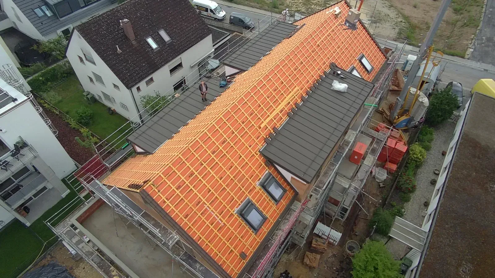 Luftaufnahme eines Hausdaches, das repariert wird; an einer Seite ist es mit orangefarbenem Dachmaterial gedeckt und ein Gerüst aufgebaut.