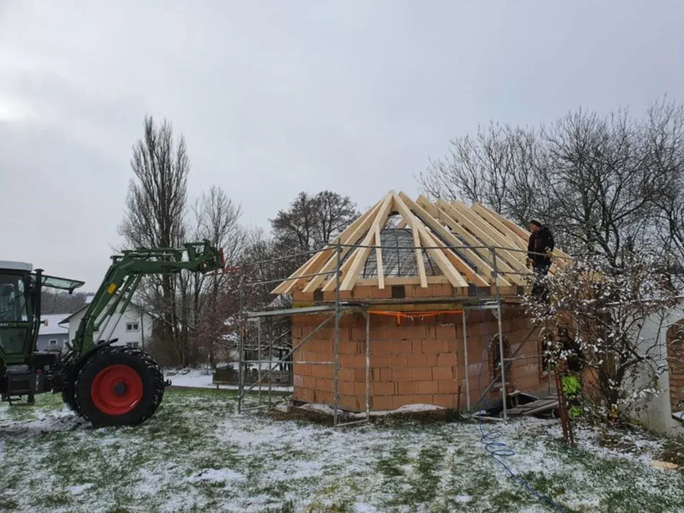 In einem verschneiten Hof befindet sich ein im Bau befindlicher Holzschuppen; auf dem Dachgerüst stehen ein Traktor und ein Arbeiter.