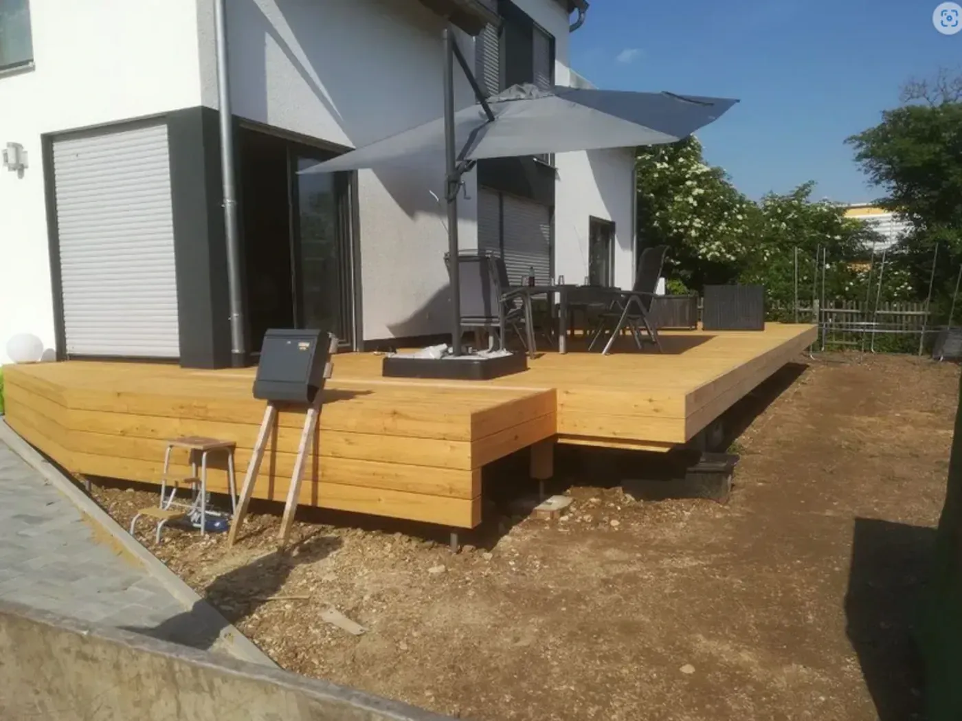 Eine Holzterrasse neben einem weißen Haus mit schwarzem Sonnenschirm, Stühlen und einem kleinen Grill im Garten