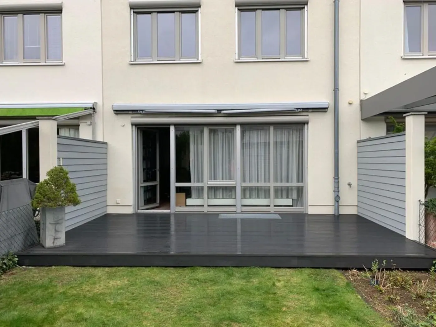 Hinterhofterrasse mit schwarzem Holzdeck, Glasschiebetüren, grauem Zaun und grünem Rasen hinter einem Haus