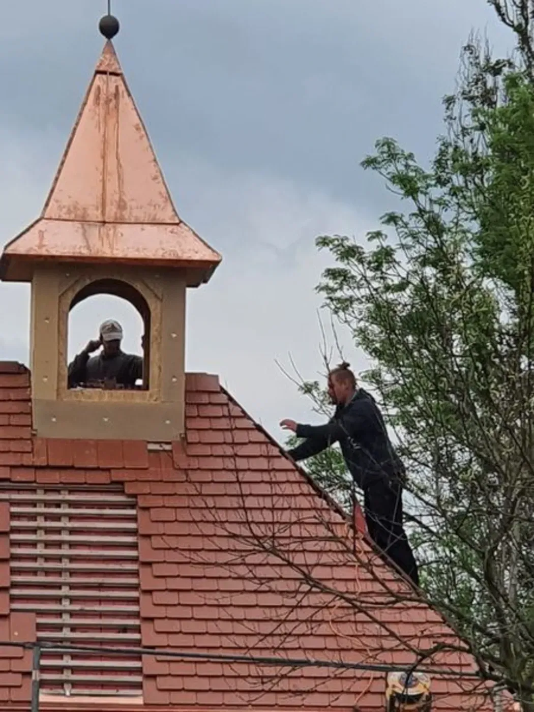 Eine Person auf einem Dach neben einem kleinen Glockenturm stutzt Äste in der Nähe der Dachspitze.