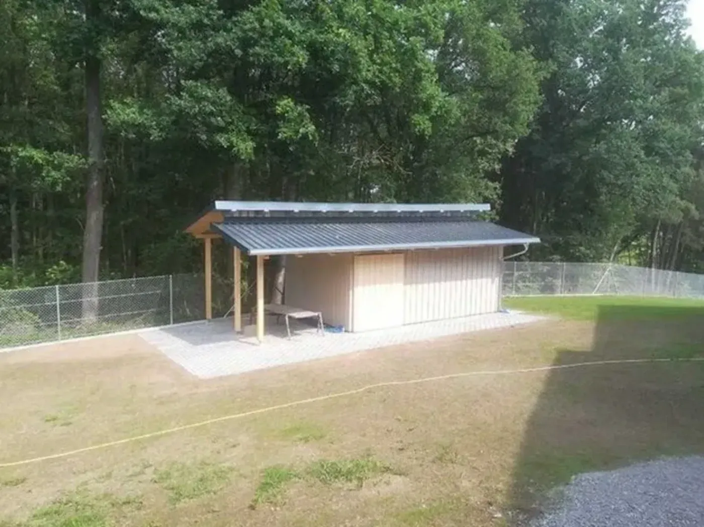 Kleiner Schuppen mit Solardach in einem grasbewachsenen Hof neben einem Waldstück und einem Zaun