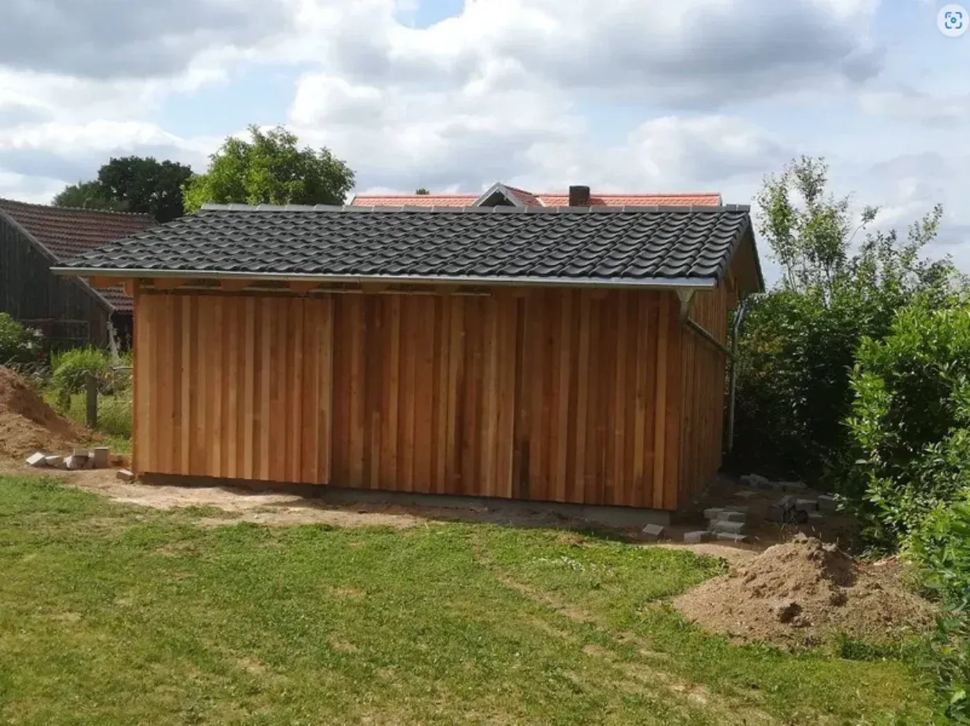 Ein Holzschuppen mit dunklem Ziegeldach an einem grasbewachsenen Hang, umgeben von Bäumen und einem bewölkten Himmel.