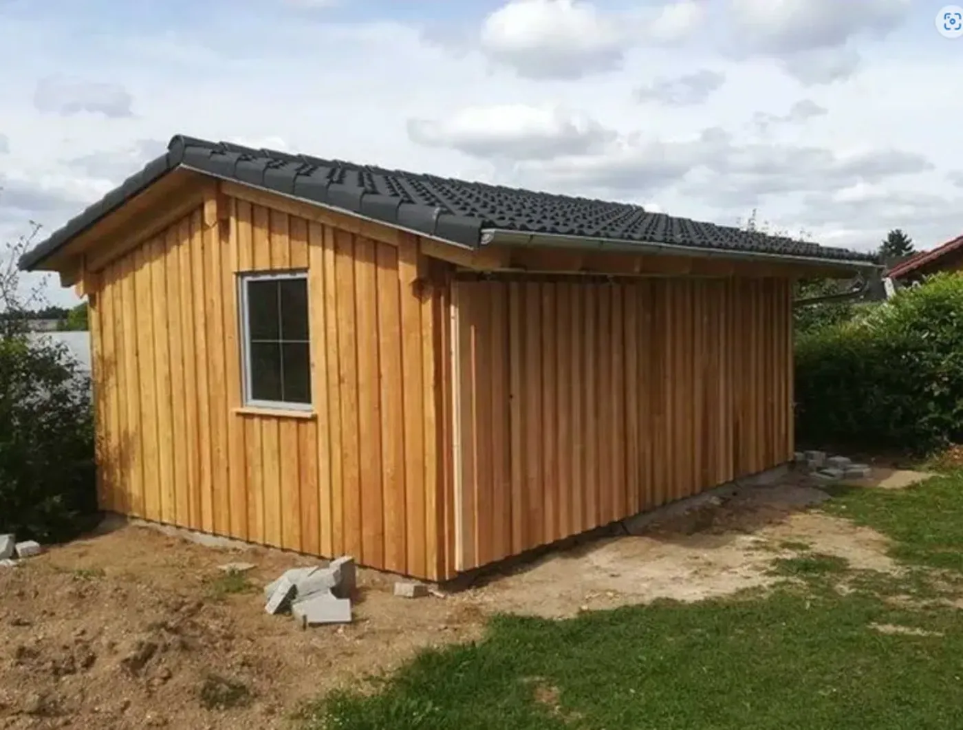 Kleiner Holzschuppen mit dunklem Dach auf einem mit Erde und Gras bedeckten Hof unter bewölktem Himmel