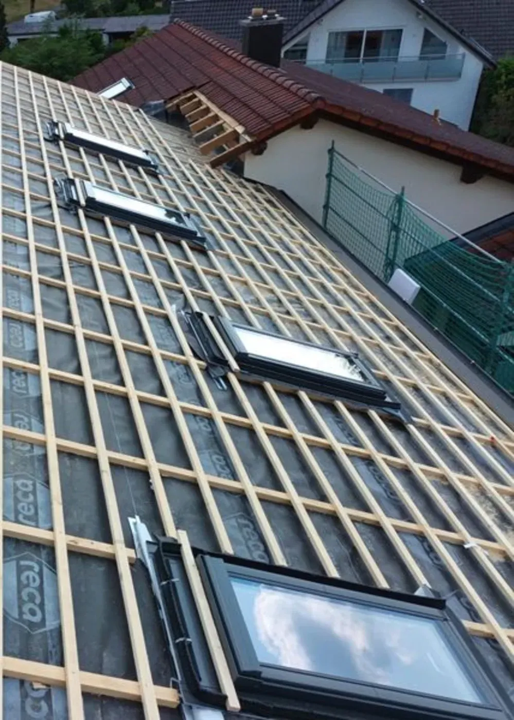 Ein Dach wird renoviert, wobei die Holzkonstruktion und die Oberlichter sichtbar sind, daneben ein Haus mit rotem Ziegeldach.
