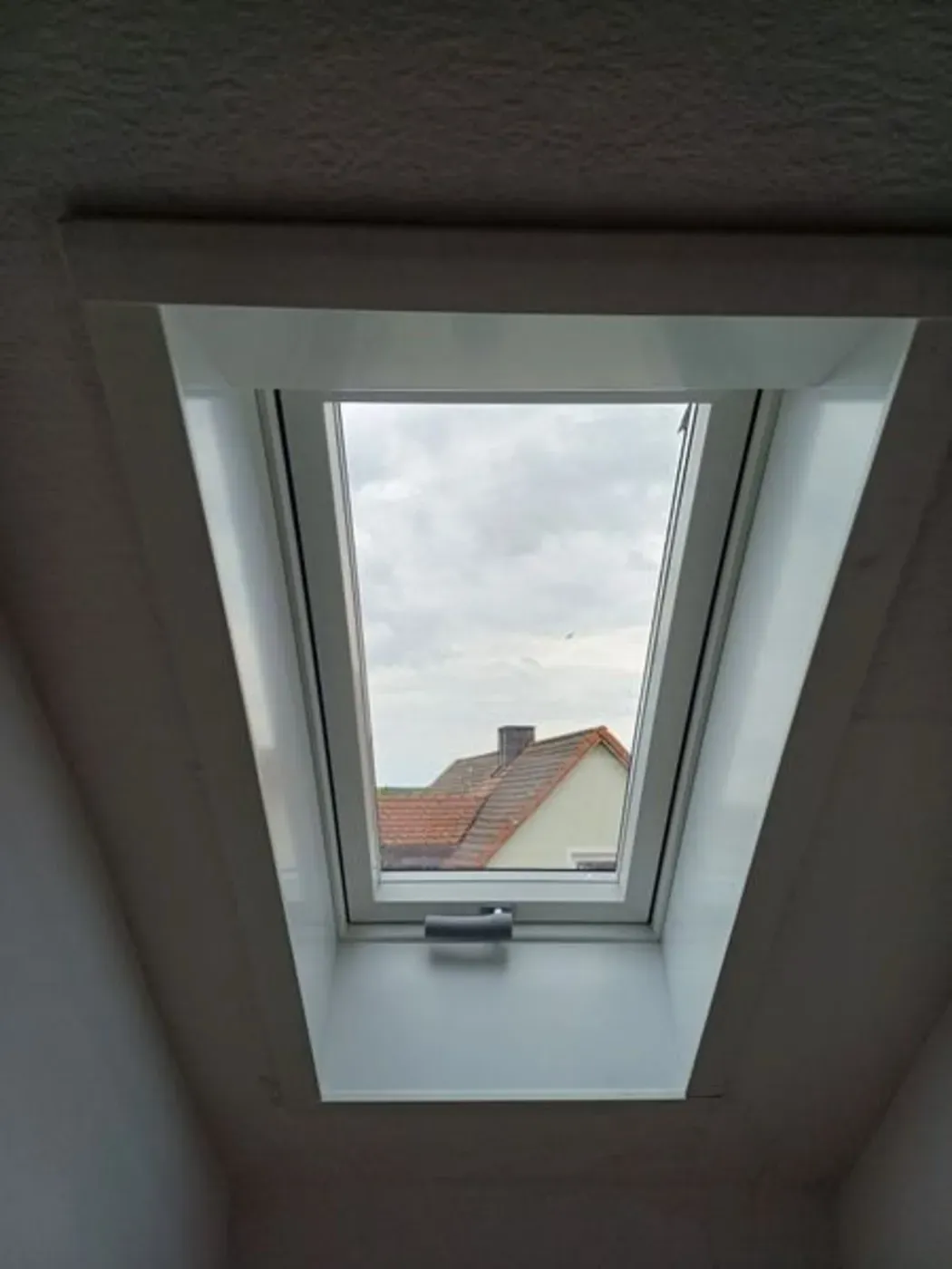 Ein Dachfenster in der Dachschräge ist geöffnet und gibt den Blick auf den bewölkten Himmel und die Dächer draußen frei.