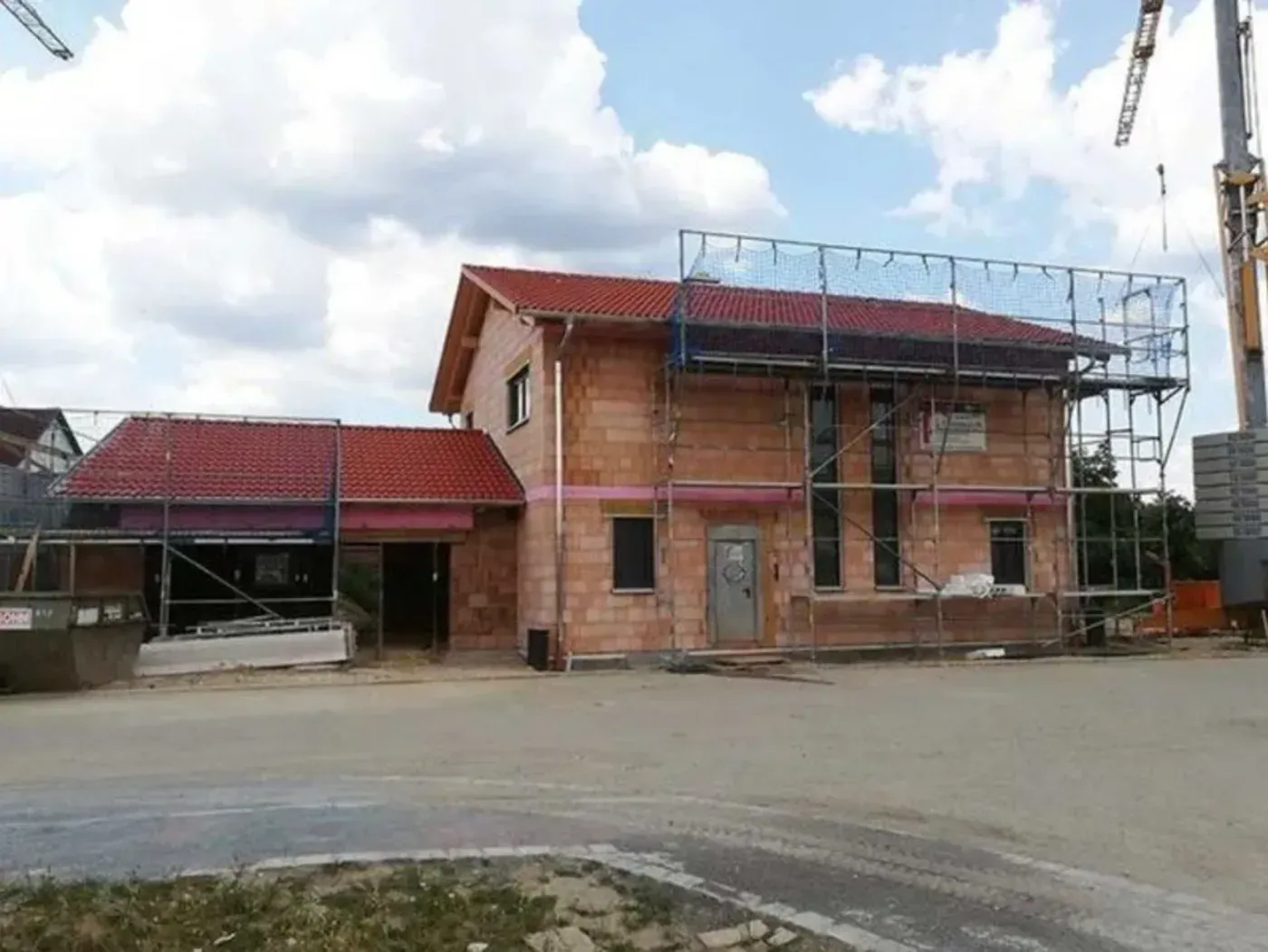 Ein teilweise fertiggestelltes Backsteinhaus mit rotem Dach und Gerüst neben einer gepflasterten Straße unter einem bewölkten Himmel