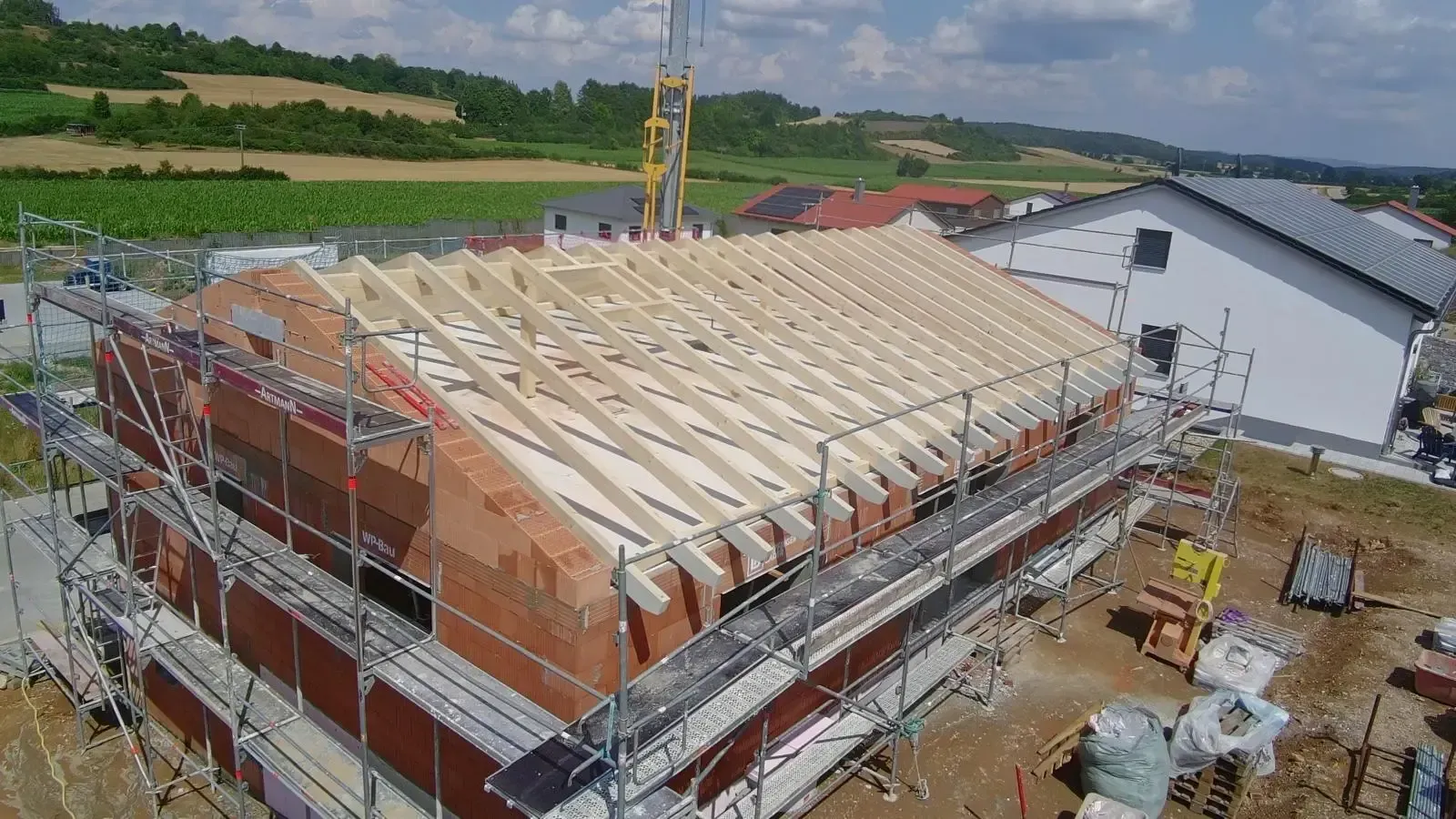 Baustelle mit teilweise errichtetem Dach, Gerüst, Kran und Arbeitern neben einem weißen Gebäude.