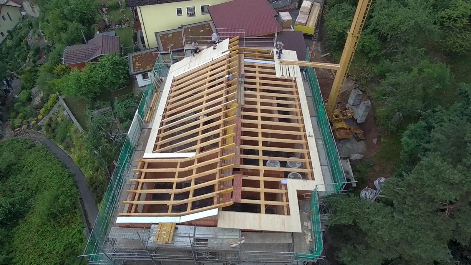 Luftaufnahme eines im Bau befindlichen Hausdaches mit freiliegenden Holzsparren und umliegenden Bäumen.