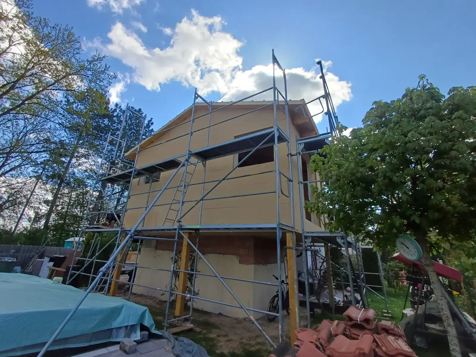 Haus im Bau mit Gerüst, beigefarbener Fassade und Bäumen unter blauem Himmel