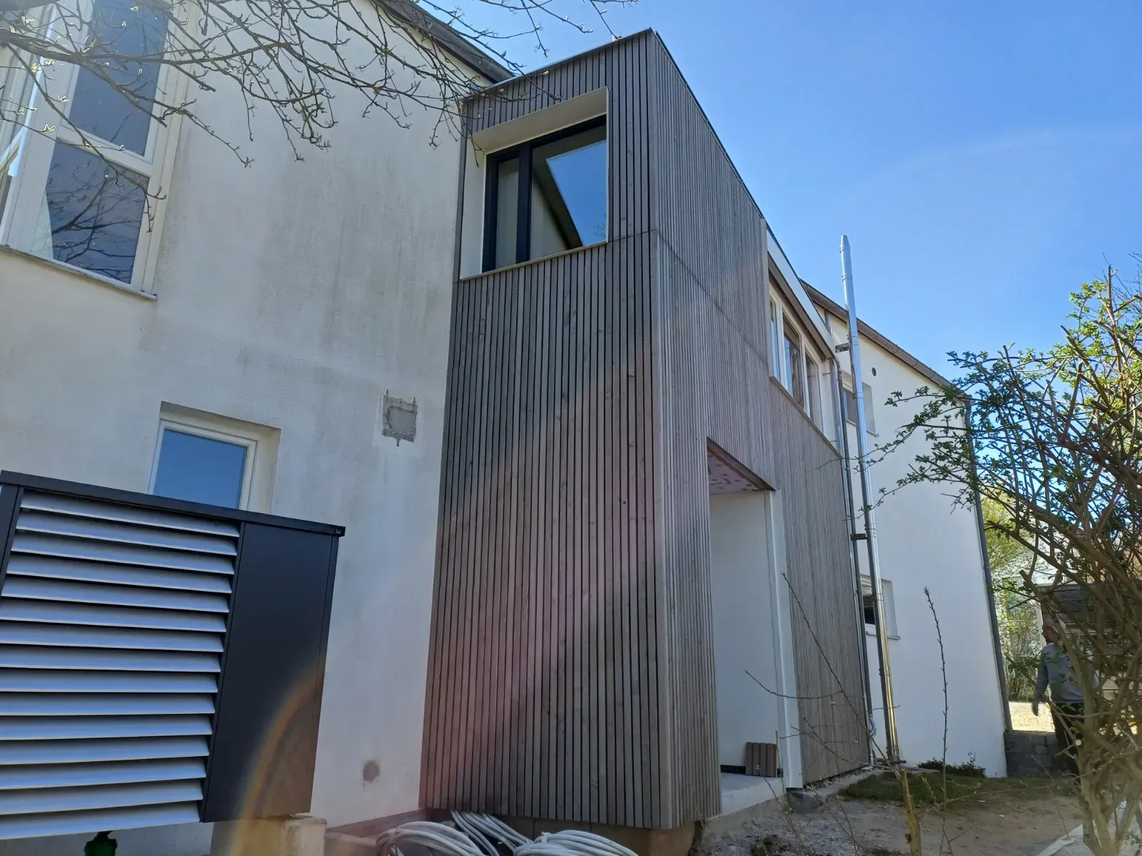 Moderne Gebäudefassade mit grauen vertikalen Lamellen und kleinen Fenstern neben einem weißen Haus unter blauem Himmel