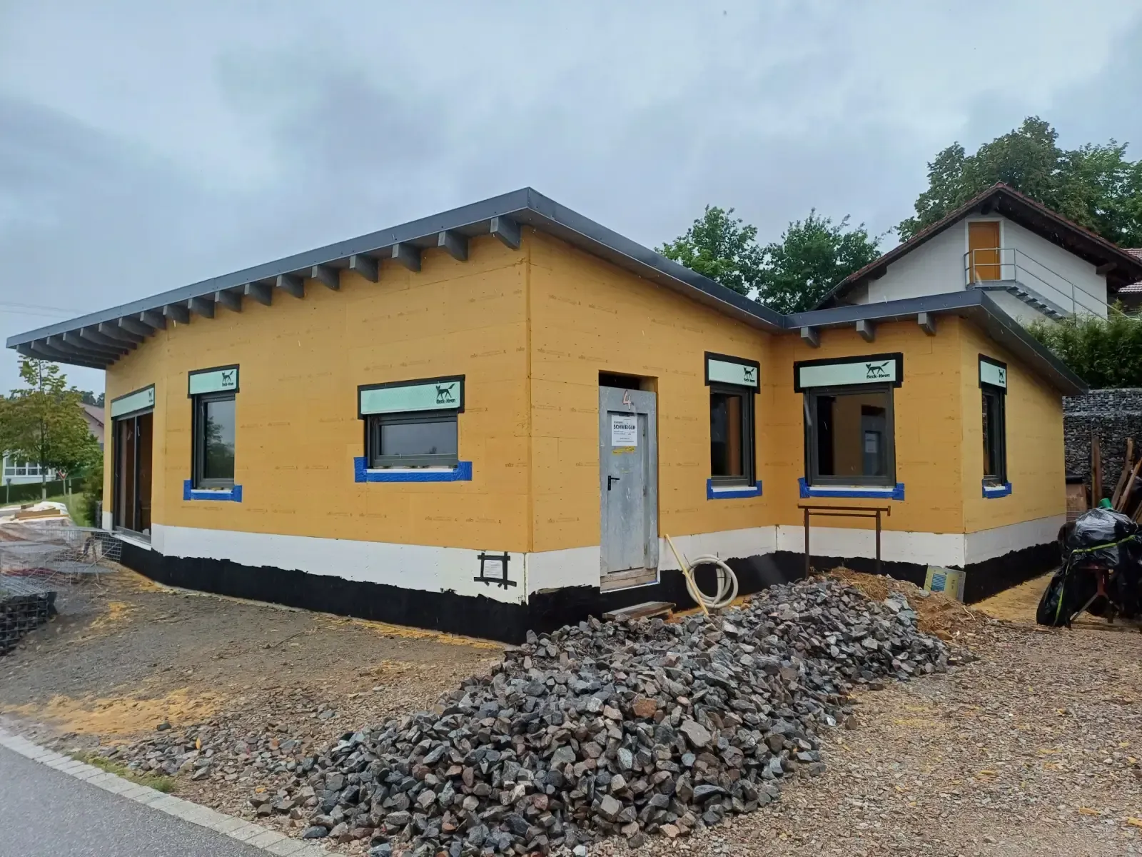 Gelbes, eingeschossiges Haus im Bau mit weißem Sockel und Kieshaufen davor.