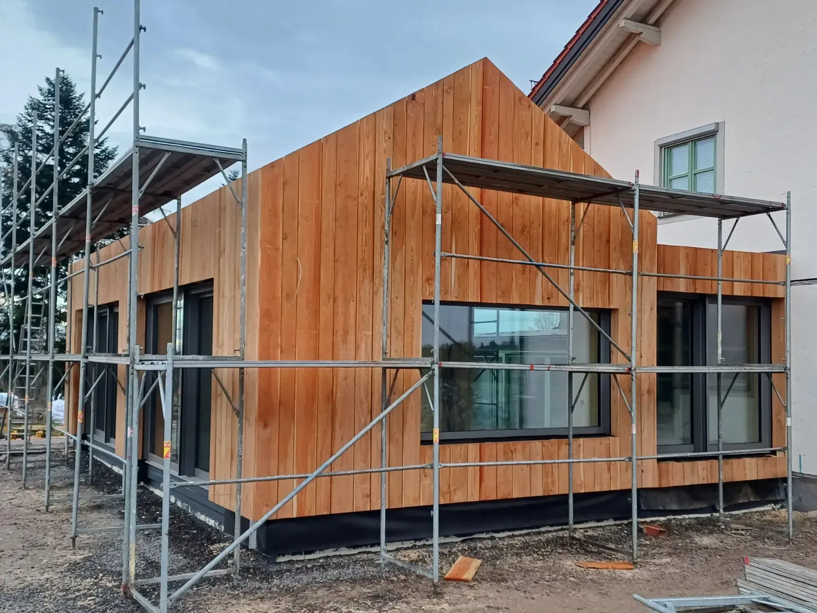 Die im Bau befindliche, mit Holz verkleidete Hausfassade ist mit einem Gerüst versehen und die Fenster sind teilweise eingebaut.